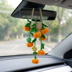 Op de afbeelding: Een gehaakt hangend auto-accessoire met een klein beige mandje met daarin verschillende oranje pompoenen met groene steeltjes en bladeren. De pompoenen zitten vast aan lange groene ranken die uit het mandje hangen, dat aan twee beige bandjes is opgehangen.