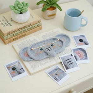 May include: A gray crochet elephant-shaped eye mask with gold-rimmed glasses resting on an open book. The mask has pink ear details and black stitched features. Miniature crochet plants and a blue mug are also present.