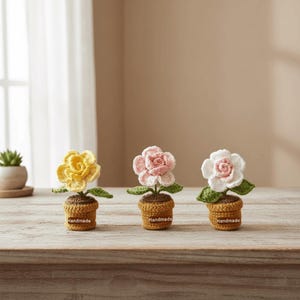 May include: Three handmade crochet flowers in small pots. The flowers are yellow, pink, and white, with green leaves. The pots are brown and have "Handmade" labels. The flowers are arranged on a wooden surface.