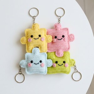 May include: Four colorful crochet puzzle piece keychains. Each keychain is a different color: yellow, pink, blue, and green. They have smiling faces with rosy cheeks and are attached to silver keyrings.