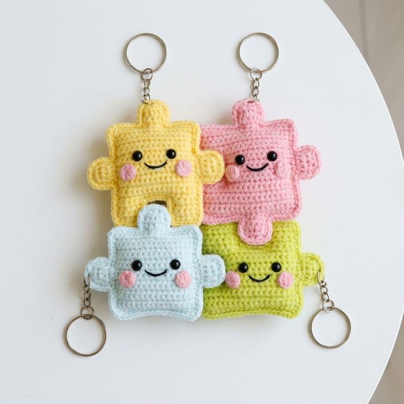 May include: Four colorful crochet puzzle piece keychains. Each keychain is a different color: yellow, pink, blue, and green. They have smiling faces with rosy cheeks and are attached to silver keyrings.