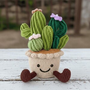 Op de afbeelding: Een gehaakte cactusopstelling in een beige pot met een lachend gezicht en bruine voetjes. De cactussen zijn in verschillende groentinten met roze en paarse bloemaccenten. De pot heeft een geschulpte rand.
