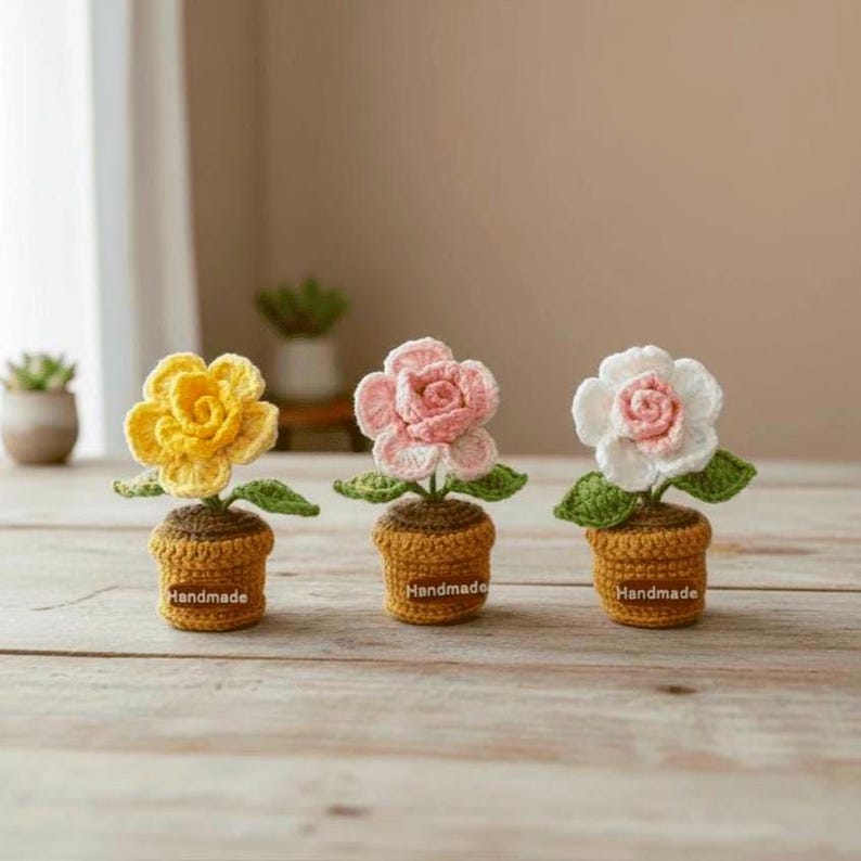May include: Three handmade crochet flowers in pots. The flowers are yellow, pink, and white, with green leaves. The pots are brown and have the word "Handmade" on them. The flowers are on a wooden surface.