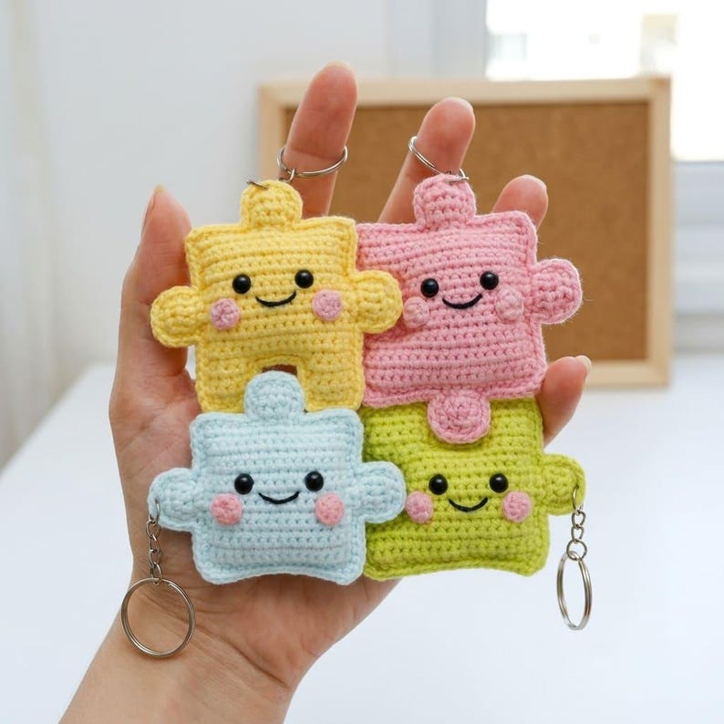 May include: Four crocheted puzzle piece keychains in yellow, pink, light blue, and green. Each piece has a smiling face with rosy cheeks and a silver keyring attached. The keychains are handmade and have a textured appearance.