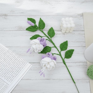 May include: A handcrafted crochet fuchsia flower stem with white and purple blossoms and green leaves. The stem is lying on a white wooden surface, with a white candle and a ball of green yarn in the background.