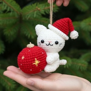 Può includere: Un ornamento natalizio fatto a mano a forma di gatto bianco all'uncinetto, con un cappello da Babbo Natale rosso, che tiene una palla di Natale rossa con dettagli a stella gialla. Sospeso da uno spago naturale.