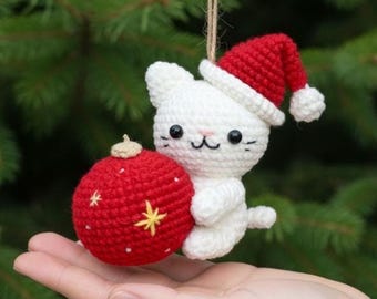 May include: A handmade white crochet cat ornament wearing a red Santa hat, holding a red Christmas ball with yellow star details. The ornament is suspended by a natural twine string, perfect for holiday decor.