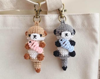 May include: Two crochet otter keychains with bronze-toned lobster clasps. One otter is light brown with a white belly and holds a small pink fish. The other is gray with a white belly, black paws, and holds a small light blue fish. Both have black eyes.