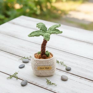 May include: A handmade crochet palm tree in a beige pot. The palm tree features a brown trunk and green leaves. The pot has a small wooden tag that reads "Hand Made". The scene includes small grey stones and greenery.