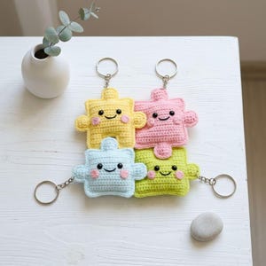 May include: Four colorful crochet puzzle piece keychains in yellow, pink, light blue, and green, each with a smiling face and attached to a silver keyring. The keychains are stacked on a white surface.