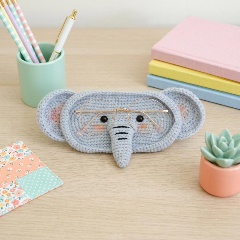 May include: A light blue crocheted elephant-shaped glasses case with pink accents. The case holds a pair of gold-rimmed glasses. The elephant has black eyes and a trunk. The case is on a light wood surface.