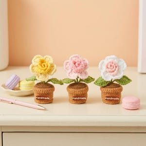 May include: Three handmade crochet flowers in small brown pots. The flowers are yellow, pink, and white, with green leaves. Each pot has a "Handmade" label. Macarons and a pen are in the background.
