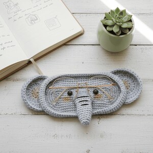 May include: A light gray, crocheted elephant-shaped glasses case with gold-rimmed glasses inside. The case has large ears and a trunk. A succulent plant in a small green pot and an open notebook are in the background.