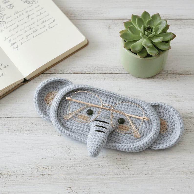 May include: A light gray crocheted elephant-shaped glasses holder with round eyeglasses. The holder has beige accents for the ears and eyes, and a small trunk. A succulent plant in a green pot and a notebook are in the background.
