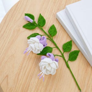 May include: A handcrafted crochet fuchsia flower stem featuring two fully bloomed white and light purple flowers, one small purple bud, and several textured green leaves attached to a thin, flexible green stem made of yarn.