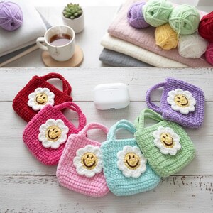 K&ouml;nnte beinhalten: Eine Sammlung farbenfroher, geh&auml;kelter Mini-Tragetaschen, jede mit einem l&auml;chelnden G&auml;nsebl&uuml;mchen-Applikation verziert. Die Taschen sind in den Farbt&ouml;nen Rot, Rosa, Lila, Blau und Gr&uuml;n gehalten. Das Bild enth&auml;lt auch Wollkn&auml;uel, eine Tasse Tee und eine kleine Sukkulente.