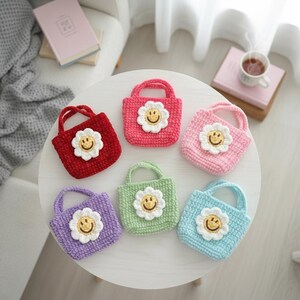K&ouml;nnte beinhalten: Sechs kleine, geh&auml;kelte Tragetaschen in verschiedenen Farben: Rot, Rosa, Hellrosa, Gr&uuml;n, Lila und Blau. Jede Tasche hat eine wei&szlig;e G&auml;nsebl&uuml;mchen-Applikation mit gelbem Smiley-Gesicht. Die Taschen haben kleine Griffe.