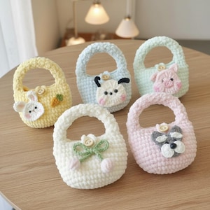 May include: A collection of five handmade crochet bags in pastel colors. Each bag features a unique animal or floral design, including a bunny, cow, pig, dog, and flowers. The bags have a rounded shape with a handle.