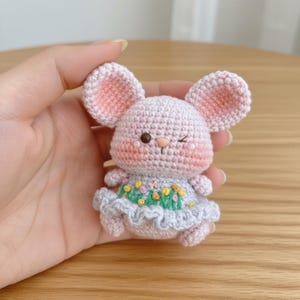 May include: A handmade crochet mouse doll in light pink with large, pink-lined ears. The mouse is wearing a light blue dress with a floral design. The doll is winking and has rosy cheeks.