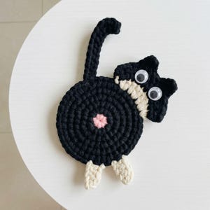 May include: A black crocheted cat coaster featuring a circular body with a pink center, a long upright tail, and two small cream-colored feet. The cat head is attached to the side, featuring two large plastic googly eyes and cream-colored accents.