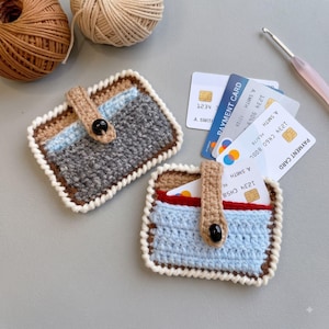 Pode incluir: Dois porta-cartões retangulares de crochê com fecho de botão. Um apresenta um design cinza e azul claro, enquanto o outro mostra um padrão azul claro e vermelho. Ambos são bordados com fio branco e contêm vários cartões de pagamento.