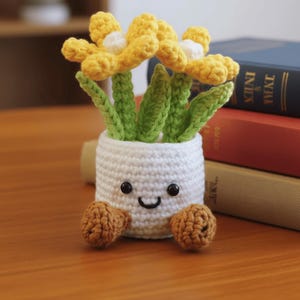 May include: A crocheted flower pot with a smiling face, brown feet, and yellow flowers with white centers. The pot is white, and the leaves are green. The handmade item is sitting on a wooden surface.