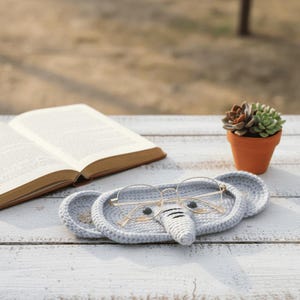 May include: A gray, crocheted elephant-shaped tray holds a pair of round eyeglasses. The tray has two large ears and a trunk. A book and a small potted succulent sit nearby on a white wooden surface.