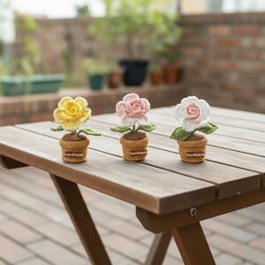 May include: Three handmade crochet flower plants in yellow, pink, and white, each in a small brown pot. The flowers have green leaves and are displayed on a wooden table. The pots have a small label that says "Handmade".