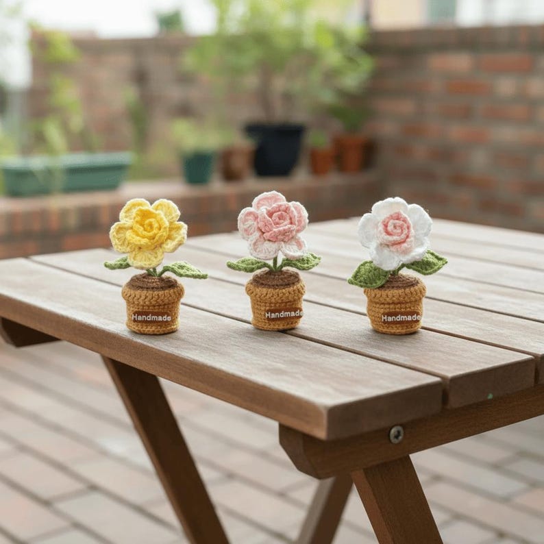 May include: Three handmade crochet flower plants in yellow, pink, and white, each in a small brown pot. The flowers have green leaves and are displayed on a wooden table. The pots have a small label that says "Handmade".