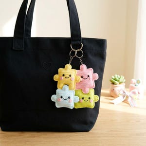 May include: A black tote bag with a small heart design, holding a keychain with four crocheted puzzle piece charms. The charms are yellow, pink, blue, and green, each with a smiling face. The keychain is attached to the bag with silver rings.