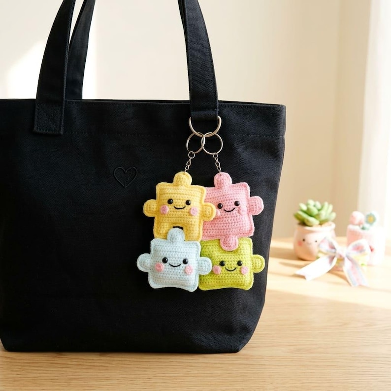 May include: A black tote bag with a small heart design, holding a keychain with four crocheted puzzle piece charms. The charms are yellow, pink, blue, and green, each with a smiling face. The keychain is attached to the bag with silver rings.