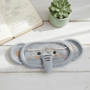 May include: A light gray, crocheted elephant-shaped glasses case with a trunk and large ears. The case holds a pair of gold-rimmed glasses. A small succulent plant in a green pot and an open notebook are in the background.