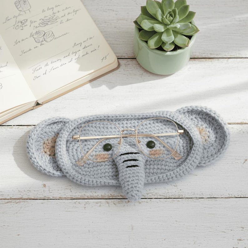 May include: A light gray crocheted elephant-shaped glasses case with gold-rimmed glasses inside. The case has tan inner ears and a trunk. The glasses case is on a white wooden surface, with a succulent plant in a green pot and an open notebook in the background.