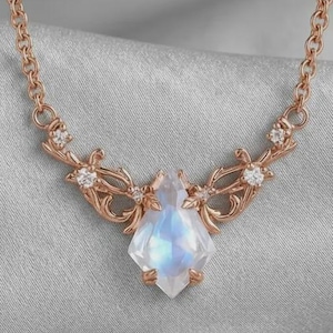 May include: Rose gold necklace featuring a central, faceted, iridescent gemstone. The pendant is flanked by ornate, floral-inspired accents with small, clear gemstones. The chain is delicate, and the necklace rests on a soft, gray surface.