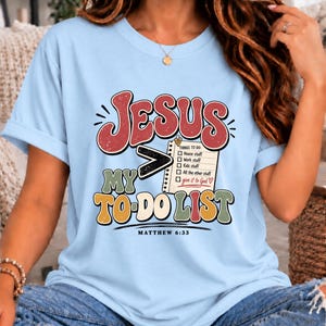 May include: A light blue t-shirt featuring a graphic with the text JESUS > MY TO-DO LIST and a checklist that reads THINGS TO DO: House stuff, Work stuff, Kids stuff, All the other stuff, give it to God, with the text MATTHEW 6:33 at the bottom.