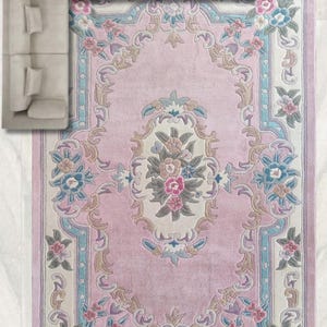 May include: A rectangular pink rug with a floral design and a light-colored border. The rug features a central floral medallion and repeating floral patterns in shades of pink, blue, and green. A beige sectional sofa is partially visible in the upper left corner.