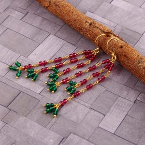 May include: Gold-toned dangle earrings with red and green beads. The earrings feature multiple strands of small, round red beads and clusters of green beads. The earrings are displayed on a wooden stick against a gray tiled background.