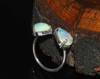 Pode incluir: Um anel de prata com dois opalas ovais. Os opalas exibem um jogo de cores com tons iridescentes de azul, verde e amarelo. A banda do anel curva-se ao redor do dedo, com cada opala definida em uma moldura de prata.