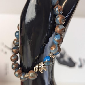 May include: A beaded bracelet with blue and brown spherical beads. The bracelet has a silver-coloured elephant charm. The beads are approximately 1.3 cm in diameter. The bracelet is displayed on a black mannequin hand.