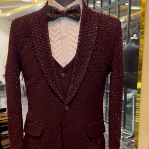 May include: A formal burgundy suit with intricate beadwork, displayed on a mannequin. The suit includes a blazer, vest, and dress shirt, paired with a patterned bow tie. The suit's design features a textured pattern and a classic lapel.