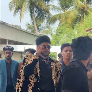 May include: A person wearing a black outfit with gold embroidery on the shoulders and sleeves, and sunglasses. The image includes palm trees and the text "MANCO.MEDIA" at the bottom.