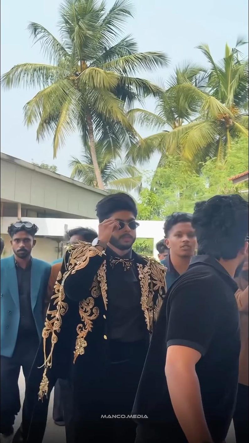 May include: A person wearing a black outfit with gold embroidery on the shoulders and sleeves, and sunglasses. The image includes palm trees and the text "MANCO.MEDIA" at the bottom.