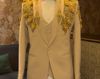 Puede incluir: Un traje color beige con bordados florales dorados en los hombros y solapas. El traje incluye una chaqueta de un solo botón, un chaleco y una pajarita negra. El traje se exhibe en un maniquí.