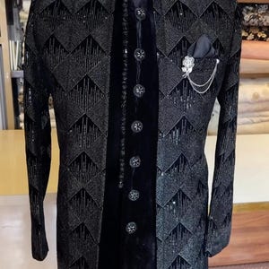 Peut inclure: Veste de c&eacute;r&eacute;monie noire avec un motif g&eacute;om&eacute;trique. La veste pr&eacute;sente un col officier, des boutons d&eacute;coratifs et une pochette avec une &eacute;pingle d&eacute;corative. La veste est pr&eacute;sent&eacute;e sur un mannequin.