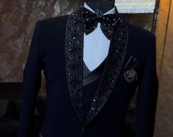 May include: A formal black tuxedo with a decorative, beaded lapel and bow tie. The tuxedo includes a white dress shirt, a black vest, and a pocket square. A small, decorative pin is on the lapel. The suit is displayed on a mannequin.