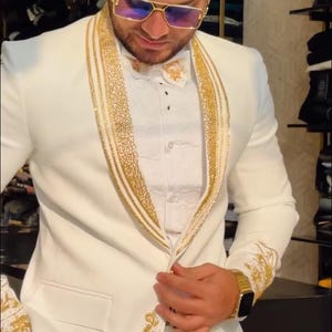 May include: A white suit with gold embroidery and trim. The suit features a notched lapel, a matching bow tie, and gold accents on the cuffs. The person is wearing gold-framed sunglasses and a gold watch. The suit is a formal wear ensemble.