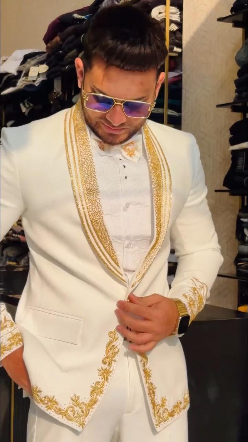 May include: A white suit with gold embroidery and trim. The suit features a notched lapel, a matching bow tie, and gold accents on the cuffs. The person is wearing gold-framed sunglasses and a gold watch. The suit is a formal wear ensemble.