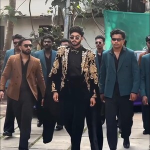 May include: A group of people walking outdoors, wearing suits and sunglasses. The central figure wears a black outfit with a gold embroidered jacket. Others wear brown and teal blazers. The image includes the text "MANCO.MEDIA".