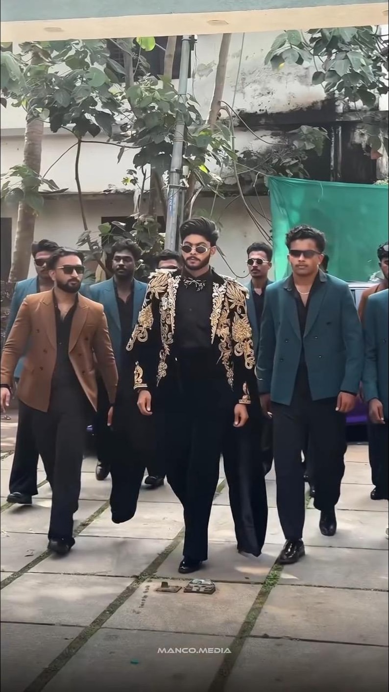 May include: A group of people walking outdoors, wearing suits and sunglasses. The central figure wears a black outfit with a gold embroidered jacket. Others wear brown and teal blazers. The image includes the text "MANCO.MEDIA".