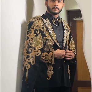 May include: Black velvet suit with gold embroidered detailing. The suit features a cape-like design with intricate floral and scrollwork patterns. The ensemble includes a black shirt and bow tie, creating a formal and elegant look.